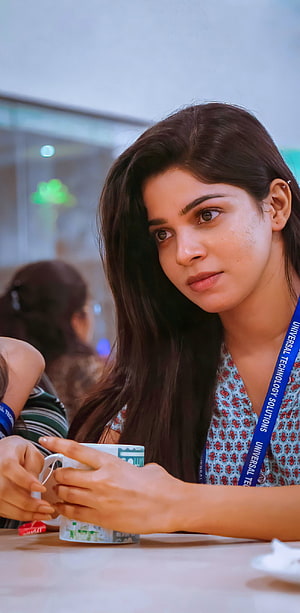 Divya Bharathi in a floral top with a blue lanyard holding a mug at a table HD phone wallpaper