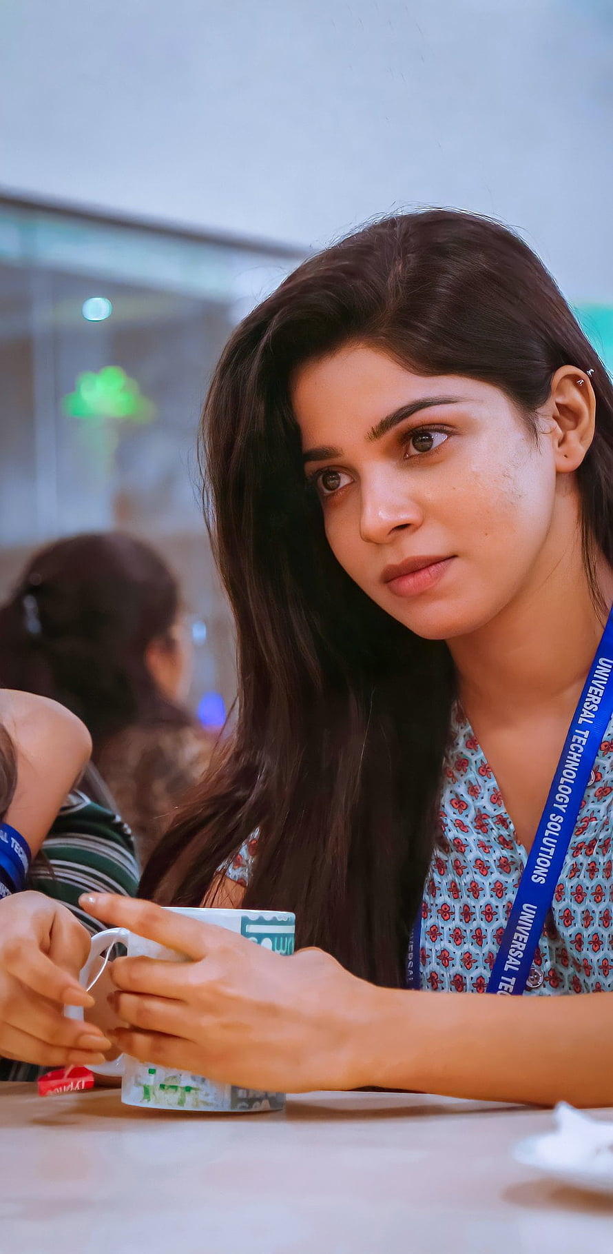 Divya Bharathi in a floral top with a blue lanyard holding a mug at a table HD phone wallpaper