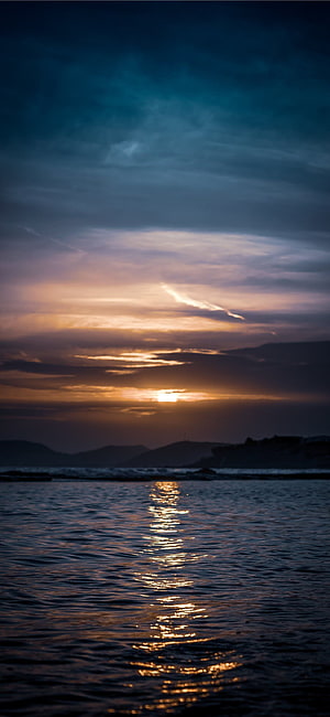 Golden sunset over calm ocean waters with distant hills and dramatic clouds HD phone wallpaper