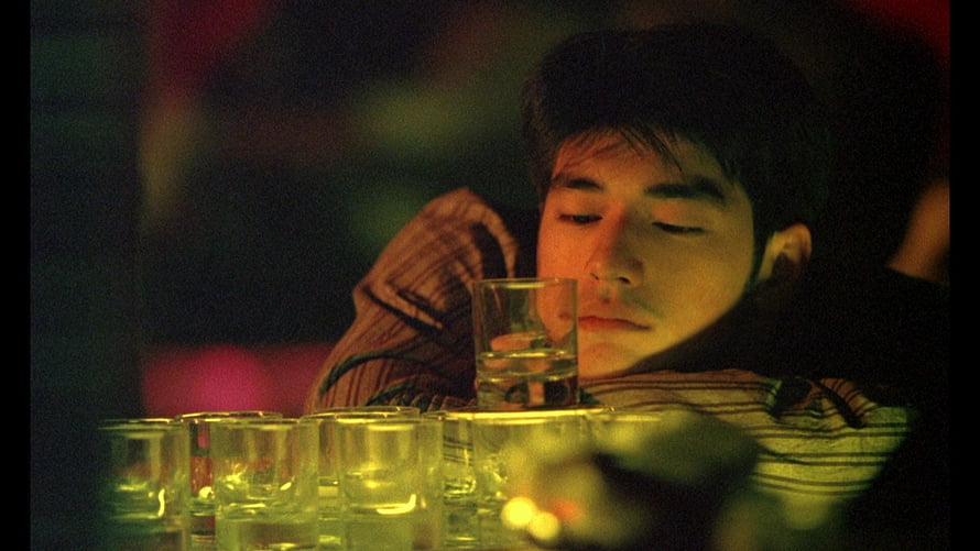 Chungking Express Film Still: Young Man Resting Amidst Rows of Small Glasses in Warm Dim Light HD wallpaper