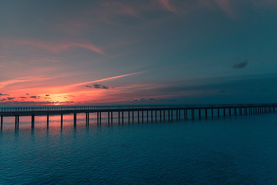 Long wooden pier extending into calm ocean waters under a vibrant sunset sky with pink and orange clouds HD wallpaper