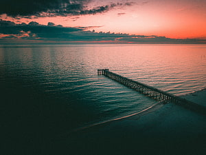 Aerial view of a wooden pier extending into a calm sea under a dramatic twilight sky with pink and orange hues HD wallpaper