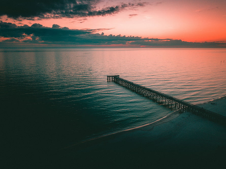 Aerial view of a wooden pier extending into a calm sea under a dramatic twilight sky with pink and orange hues HD wallpaper
