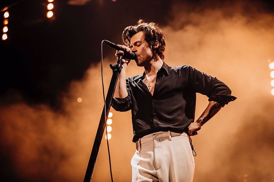 Harry Styles performing live on stage in a black shirt and white trousers with warm atmospheric lighting HD wallpaper