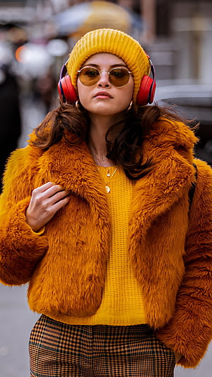 Selena Gomez in a mustard knit beanie, red headphones, and a plush amber faux fur jacket on a city street HD phone wallpaper