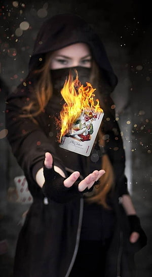 Mysterious Hooded Figure Holding a Burning Joker Playing Card in Dark Atmosphere HD phone wallpaper