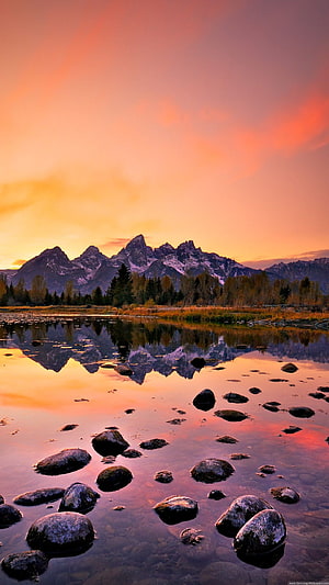 Majestic mountain range reflected in a calm river at sunset with scattered rocks and warm sky HD phone wallpaper