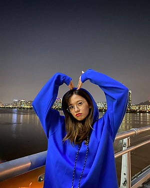 Ahn Yujin in a blue hoodie making a heart gesture against a night city skyline HD phone wallpaper