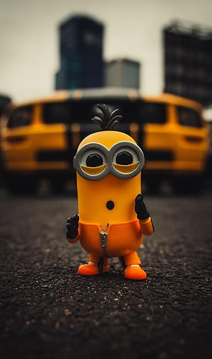 Despicable Me Minion Figurine Standing on Asphalt with Blurred Yellow Car and City Skyline Background HD phone wallpaper