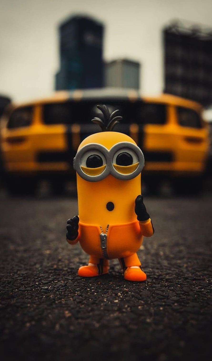 Despicable Me Minion Figurine Standing on Asphalt with Blurred Yellow Car and City Skyline Background HD phone wallpaper
