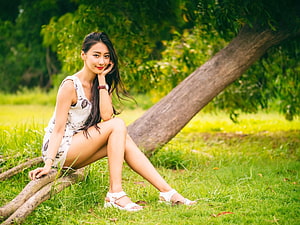 Asian woman in floral dress sitting on tree roots in a lush green park HD wallpaper