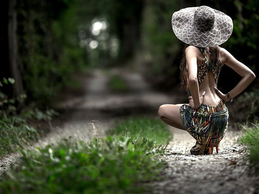Woman in patterned outfit and wide-brimmed hat crouching on a forest path HD wallpaper