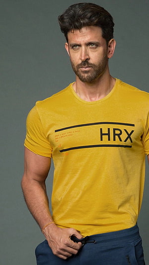 Hrithik Roshan in a yellow HRX t-shirt with navy joggers against a grey studio backdrop HD phone wallpaper