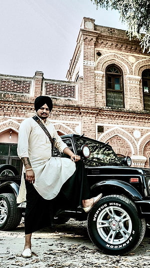 Sidhu Moose Wala in Traditional Attire Leaning Against a Black Jeep in Front of a Historic Brick Building HD phone wallpaper