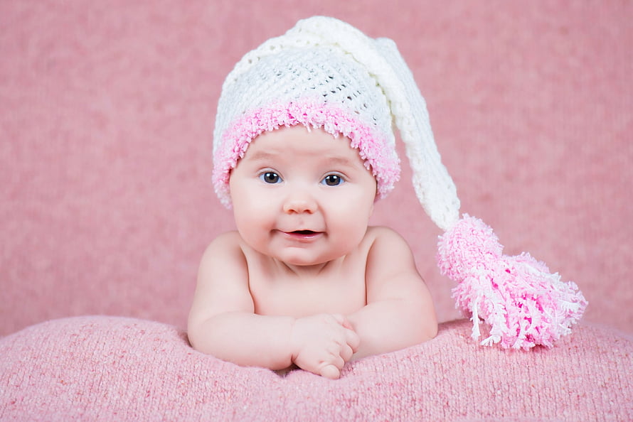 Adorable baby girl wearing a white and pink knitted hat with a tassel against a soft pink background HD wallpaper
