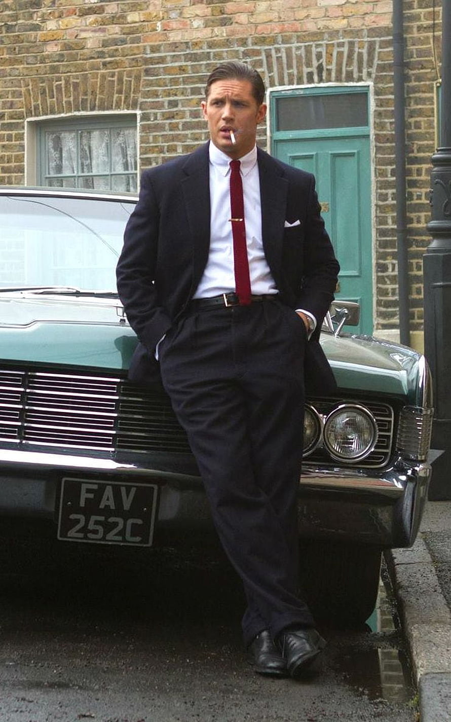 Tom Hardy in a dark suit and red tie leaning against a vintage car on a brick street HD phone wallpaper