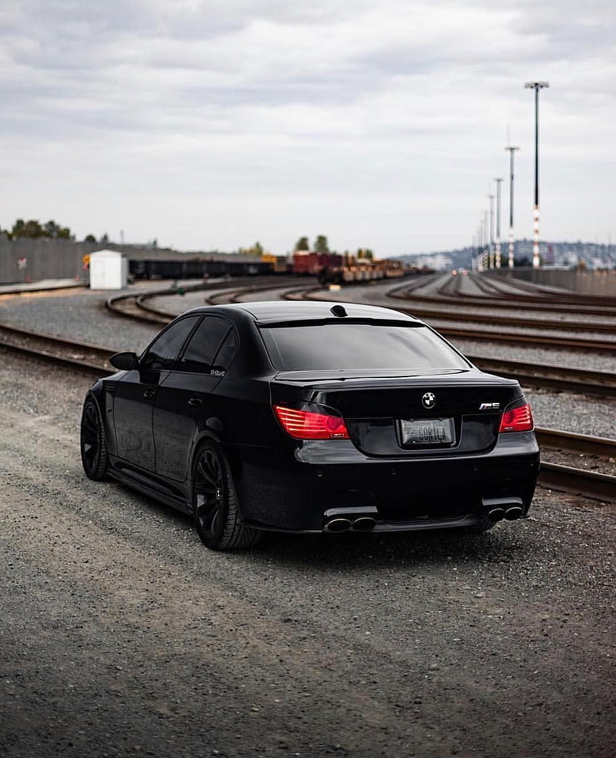 Black BMW E60 M5 Sedan Rear View Parked on Gravel Railway Tracks Under Overcast Sky HD phone wallpaper