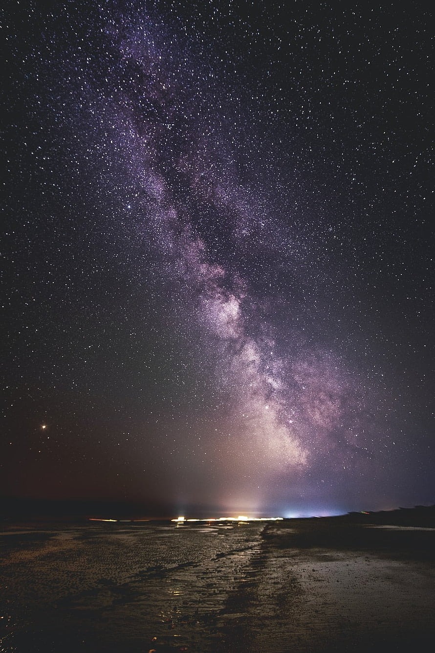 Milky Way Galaxy Arching Over a Dark Beach at Night with Distant Shoreline Lights HD phone wallpaper