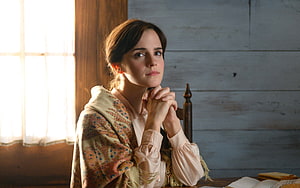 Emma Watson as Jo March in Little Women, seated at a wooden table with hands clasped in a rustic interior HD wallpaper