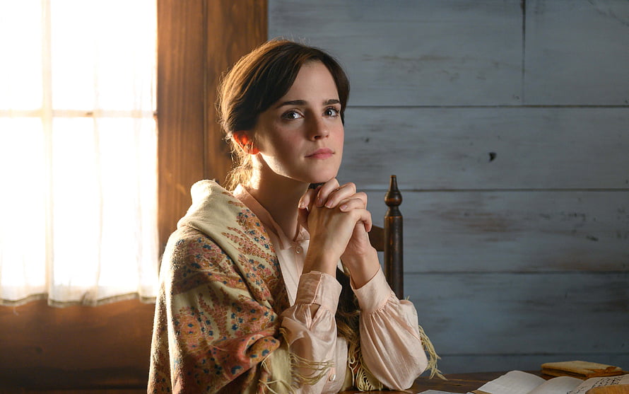 Emma Watson as Jo March in Little Women, seated at a wooden table with hands clasped in a rustic interior HD wallpaper