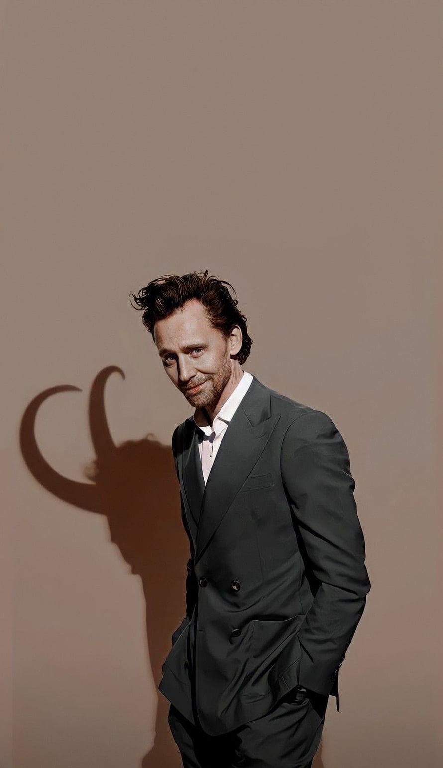 Tom Hilderton as Loki in a dark suit with a horned shadow cast on a beige wall HD phone wallpaper