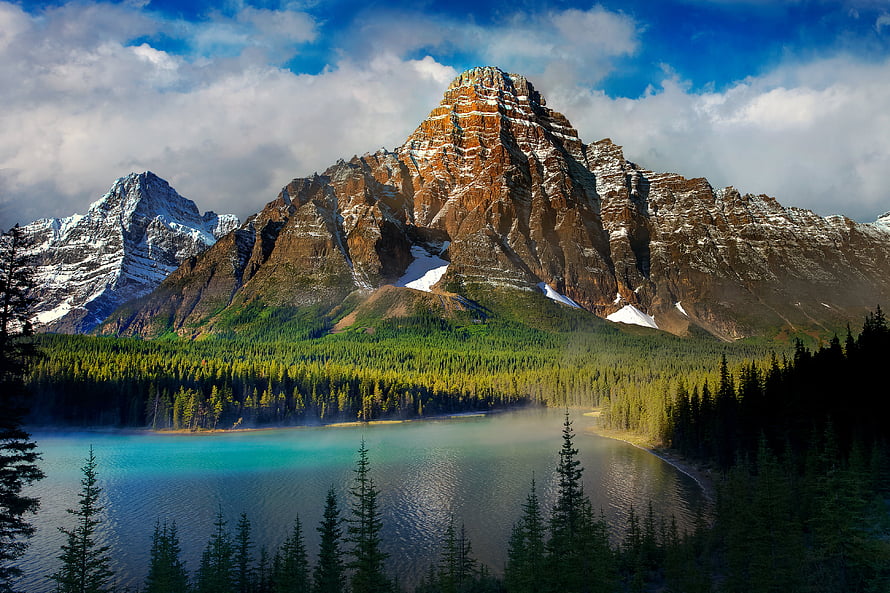 Misty turquoise alpine lake surrounded by dense evergreen forest and rugged snow-dusted mountain peaks under a dramatic sky HD wallpaper