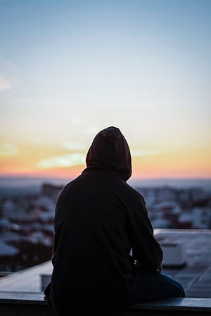 Silhouette of a hooded figure sitting on a rooftop overlooking a city at sunset HD phone wallpaper