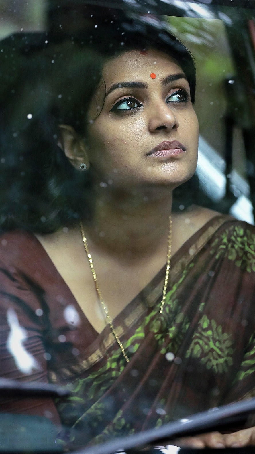 Divya Pillai in a brown and green sari with a red bindi, gazing upward through a rain-streaked window HD phone wallpaper