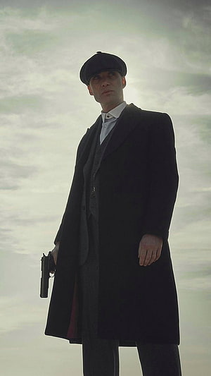 Tommy Shelby from Peaky Blinders in a long coat and flat cap holding a pistol against a cloudy sky HD phone wallpaper
