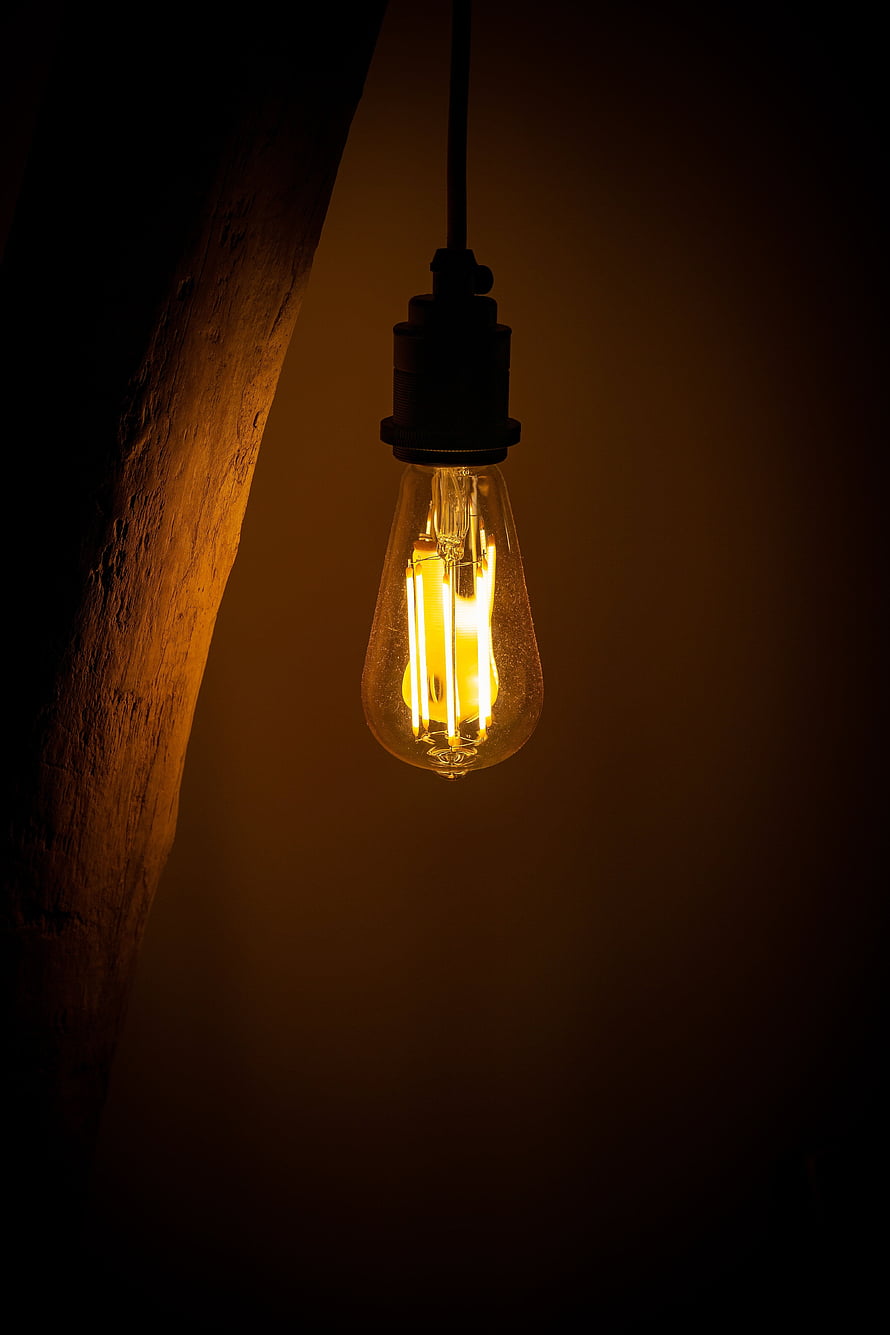 Glowing Edison filament bulb hanging in a dark room with warm amber light and rustic wood texture HD phone wallpaper