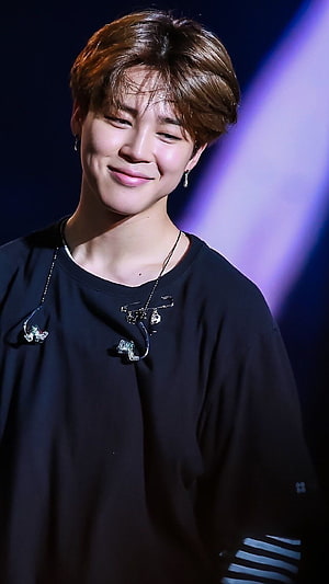 Jimin from BTS smiling on stage in a black t-shirt with in-ear monitors and silver jewelry HD phone wallpaper