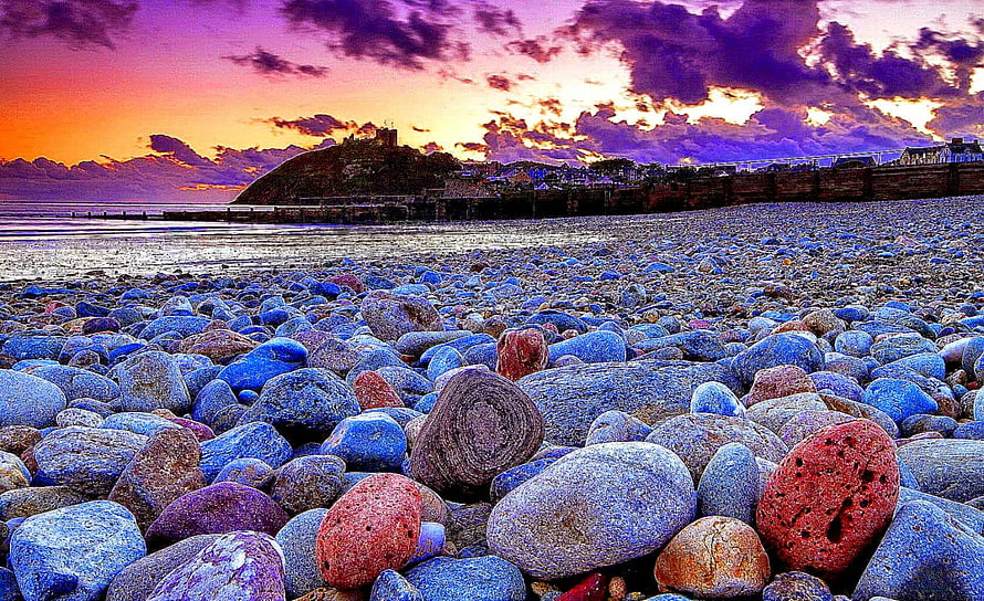 Vibrant pebble beach at sunset with dramatic purple and orange sky over coastal town HD wallpaper