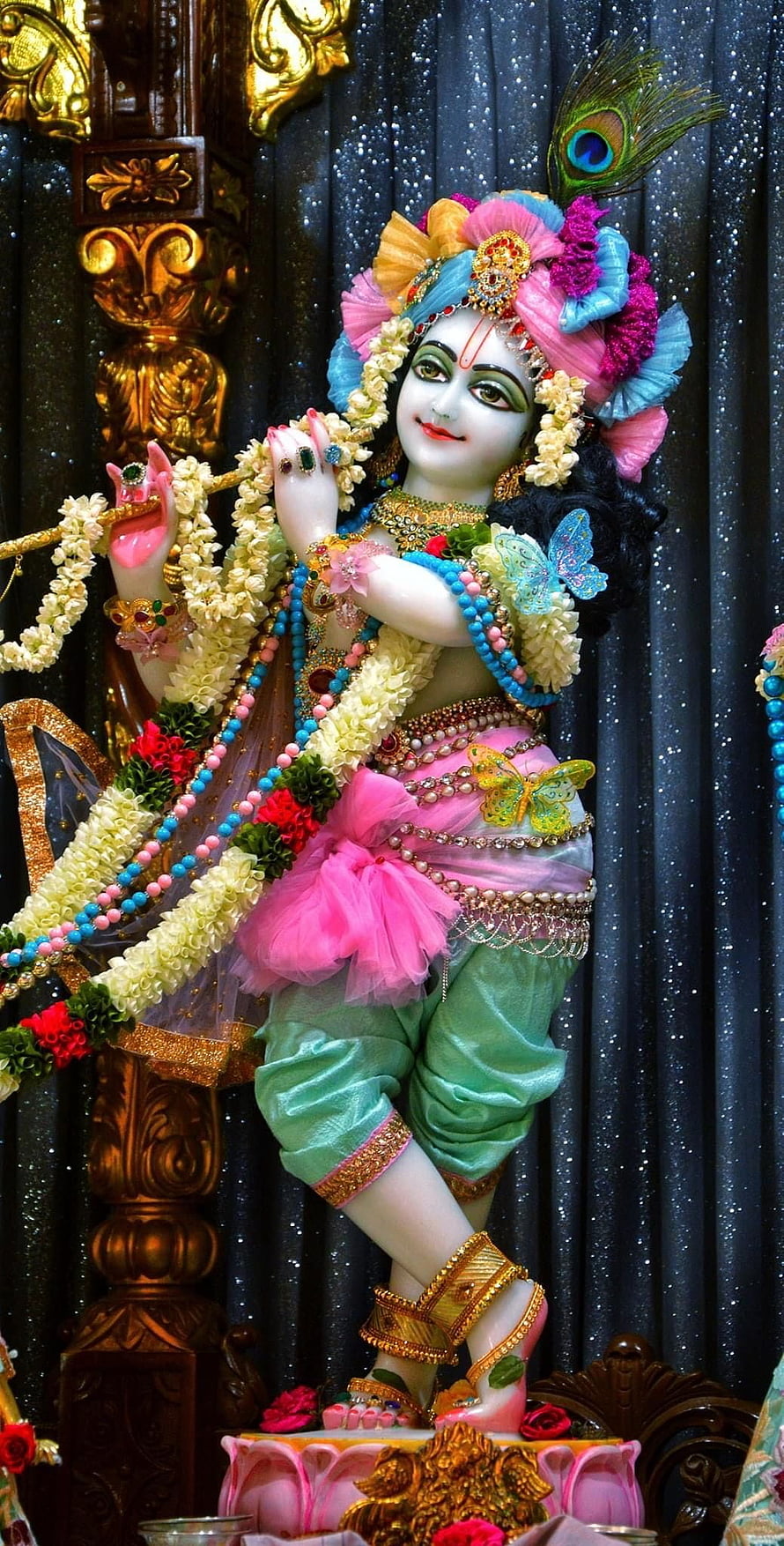Lord Krishna Murti adorned with vibrant floral garlands and peacock feather in ornate temple setting HD phone wallpaper