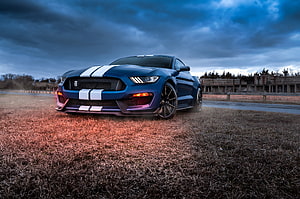 Blue Ford Mustang Shelby GT500 with white racing stripes parked on dry grass under a dramatic stormy sky HD wallpaper