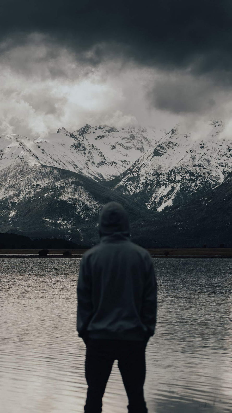 Solitary figure in a hoodie gazing at snow-capped mountains across a calm lake under a moody sky HD phone wallpaper