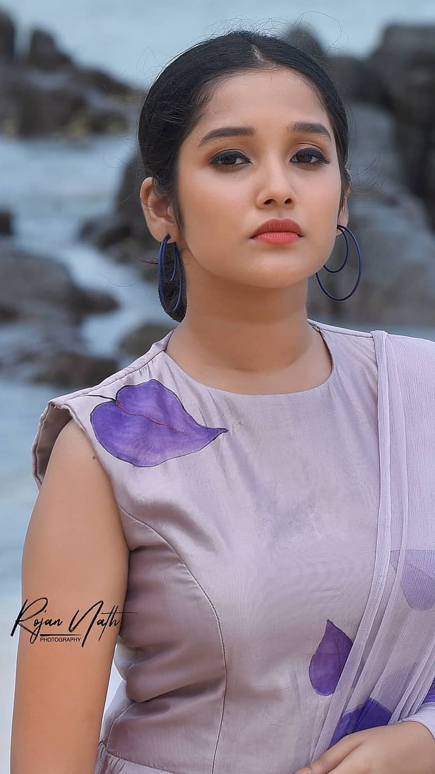Anikha Surendran in a lavender floral blouse with blue hoop earrings against a rocky coastal backdrop HD phone wallpaper