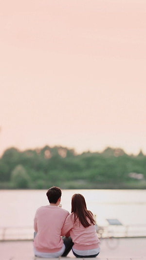 Couple in matching pink sweaters sitting by a calm lake at sunset HD phone wallpaper