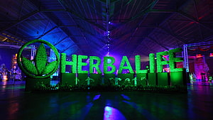 Herbalife Logo Displayed in Vibrant Green Foliage at a Large Indoor Event Venue HD wallpaper