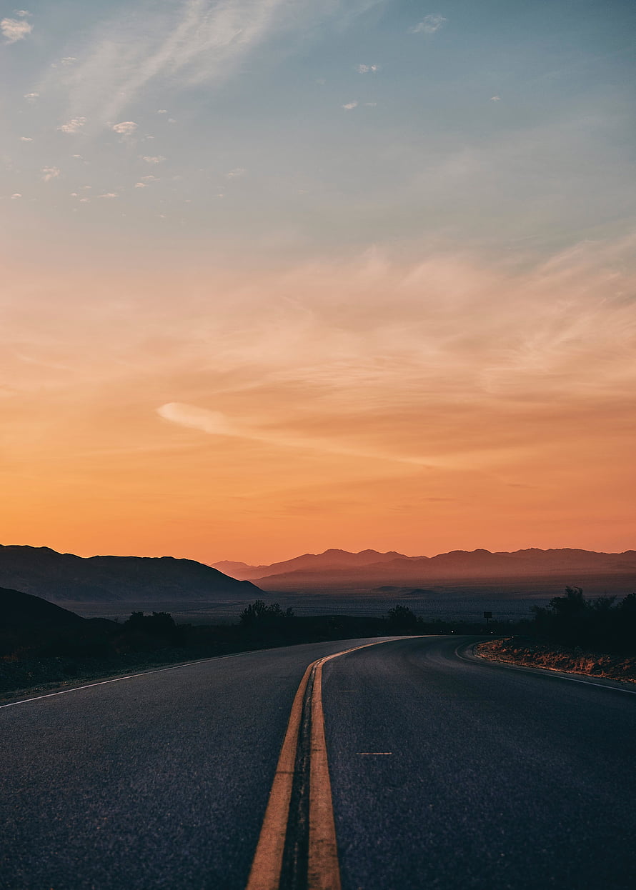 Empty Highway Stretching Toward Distant Mountains Under a Warm Orange Sunset Sky HD phone wallpaper