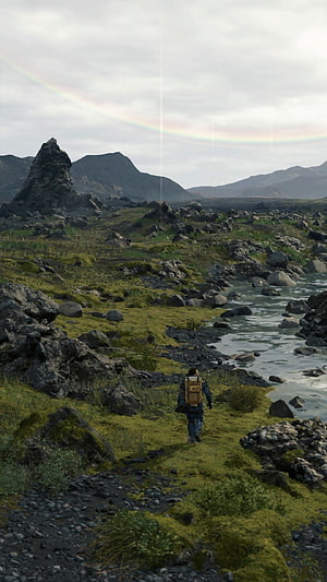 Death Stranding character walking through mossy rocky landscape with river and rainbow HD phone wallpaper