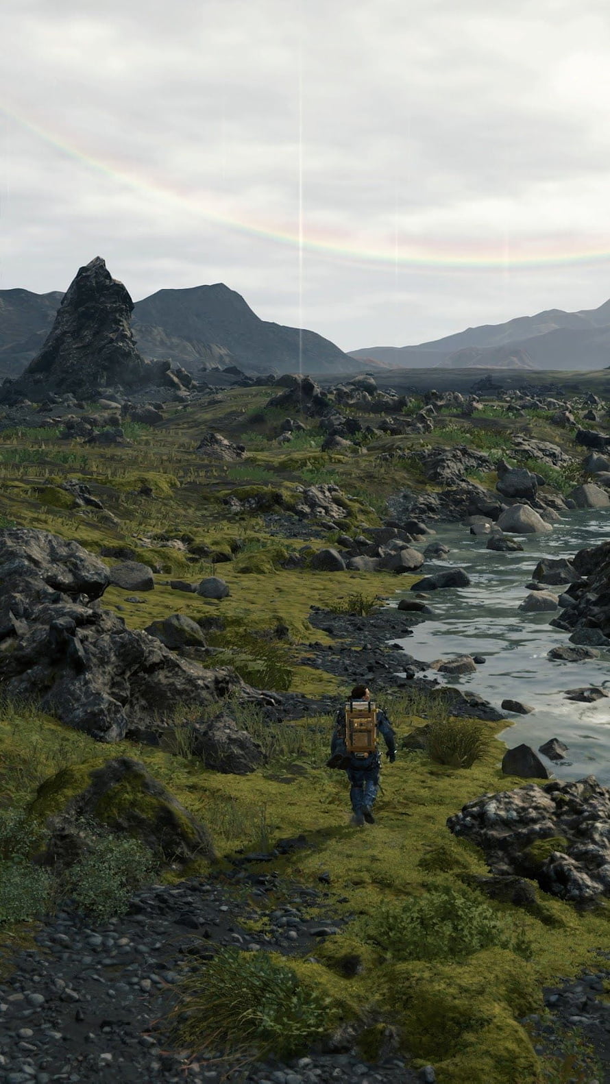 Death Stranding character walking through mossy rocky landscape with river and rainbow HD phone wallpaper