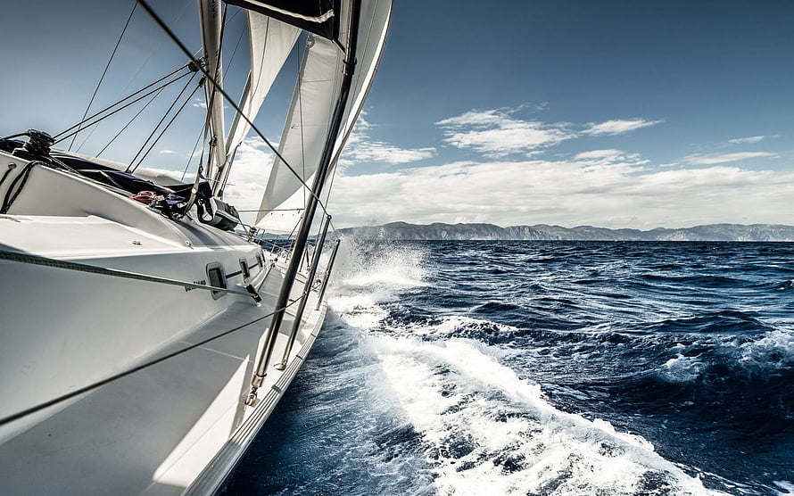 Sleek white sailboat cutting through deep blue ocean waves with distant coastline under a clear sky HD wallpaper