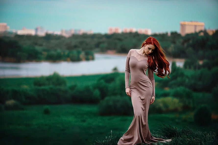 Red-haired woman in a long mauve dress standing in a green field with a river and city skyline in the background HD wallpaper