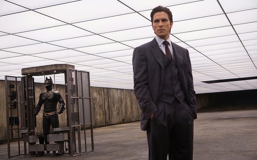 Christian Bale as Bruce Wayne in a dark suit with the Batman suit displayed in a high-tech facility HD wallpaper
