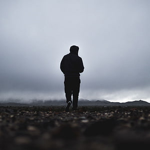 Silhouette of a person in a hoodie walking through a misty, rocky landscape under an overcast sky HD phone wallpaper