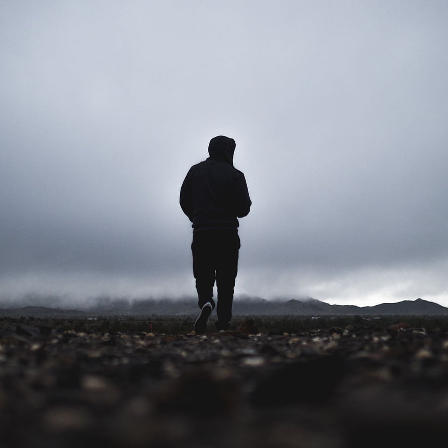 Silhouette of a person in a hoodie walking through a misty, rocky landscape under an overcast sky HD phone wallpaper