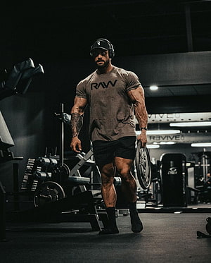 Cbum in RAW shirt walking through a dark gym holding a weight plate HD phone wallpaper