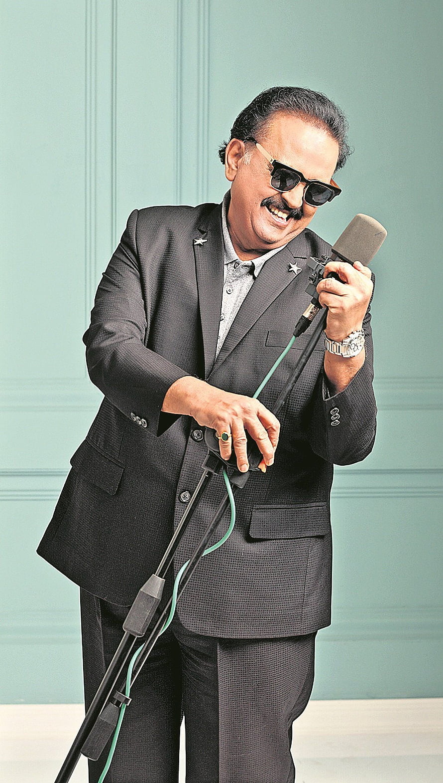 S. P. Balasubrahmanyam in a dark suit and sunglasses holding a microphone against a teal wall HD phone wallpaper