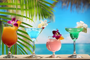 Four Colorful Tropical Cocktails Garnished With Orchids Against A Beach Backdrop HD wallpaper