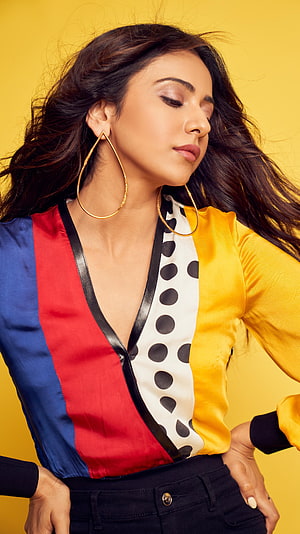 Rakul Preet Singh in a colorful striped blouse with gold hoop earrings against a yellow background HD phone wallpaper